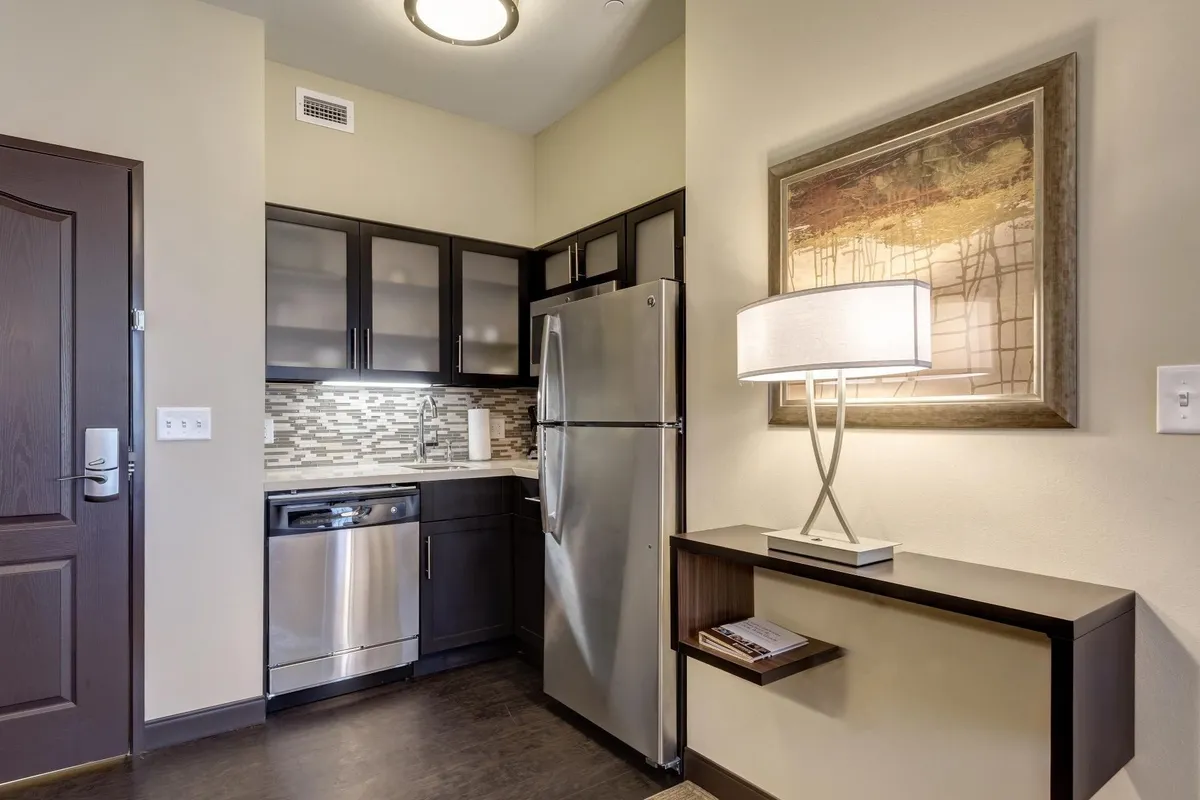 A modern studio interior featuring stainless steel appliances, sleek cabinetry, decorative artwork, and a cozy lampshade. The space exudes a functional yet inviting atmosphere ideal for cooking, relaxing, or socializing. Though the main focus is the kitchen and living area, you can imagine the versatility of the space—perfect for enjoying comfort and convenience.