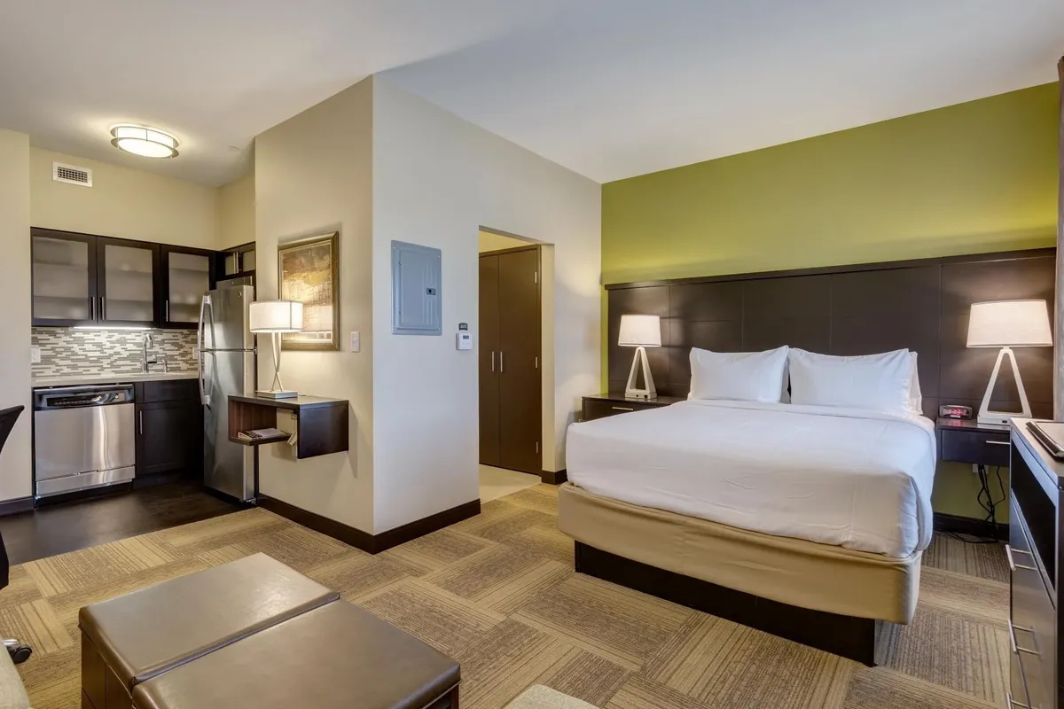 The image showcases a bright, inviting studio suite with a plush queen bed, a modern kitchenette, and a convenient work desk. The open layout is designed for comfort and flexibility, perfect for relaxing evenings, meal prep, or catching up on work. Soft lighting and tasteful décor create a cozy, welcoming atmosphere.