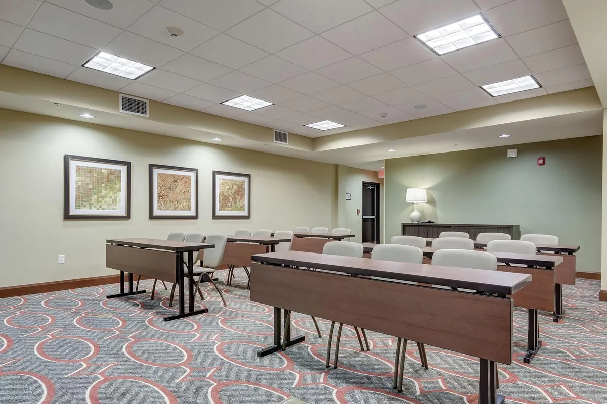 A bright and airy interior meeting space featuring rows of tables and chairs, framed artwork on the walls, and a lamp on a side table. The open-plan setup suggests a multi-functional area ideal for both work and relaxation.
