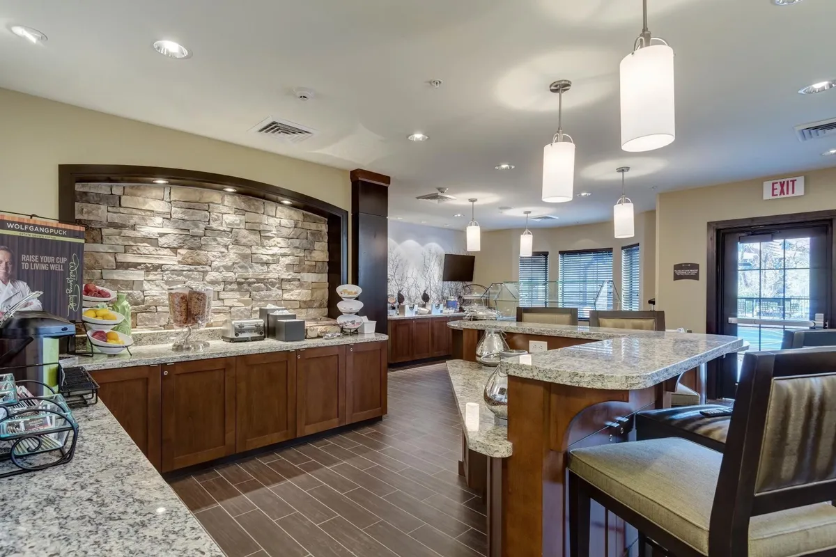 The image features a bright, inviting breakfast bar with granite countertops and a stone accent wall. A variety of food items is spread out on the bar, creating a communal atmosphere where guests can enjoy breakfast, coffee, and conversation.