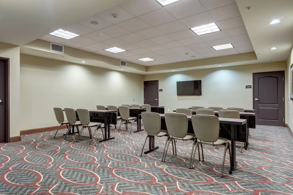 A well-lit meeting room set up in a classroom style, featuring several tables and chairs facing a mounted television at the front. The room is designed for business meetings, training sessions, or presentations, with a clean, professional atmosphere.